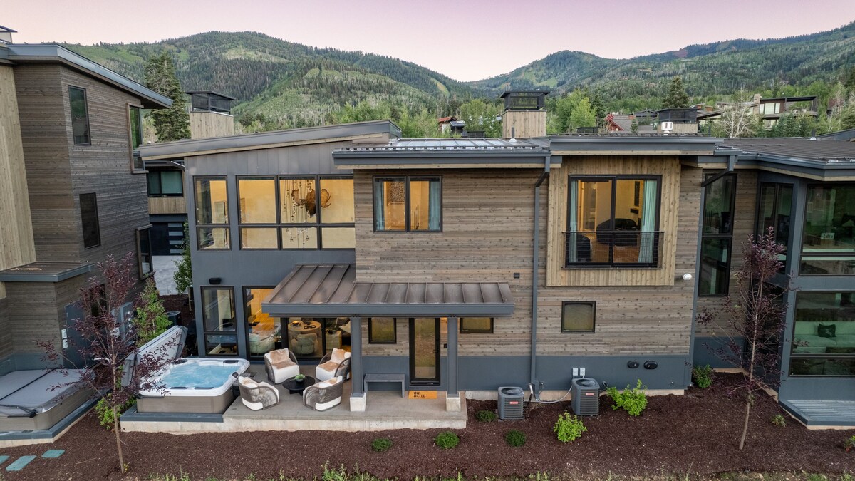 The exterior of the home reveals a modern design with a combination of wood and metal finishes. A hot tub is positioned on a patio area, accompanied by stylish outdoor seating. Lush green landscaping contrasts with the earthy tones of the structure and mountains in the background.
