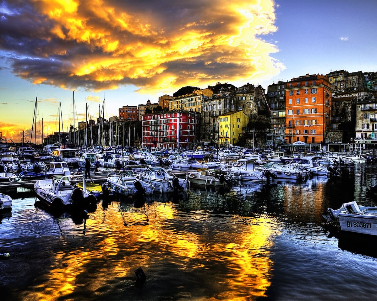 A scenic view of a harbor is presented, showcasing numerous boats gently bobbing on the water. The vibrant colors of nearby buildings reflect on the surface, while the sky is adorned with soft clouds illuminated by warm tones. The overall atmosphere conveys a tranquil coastal ambiance.