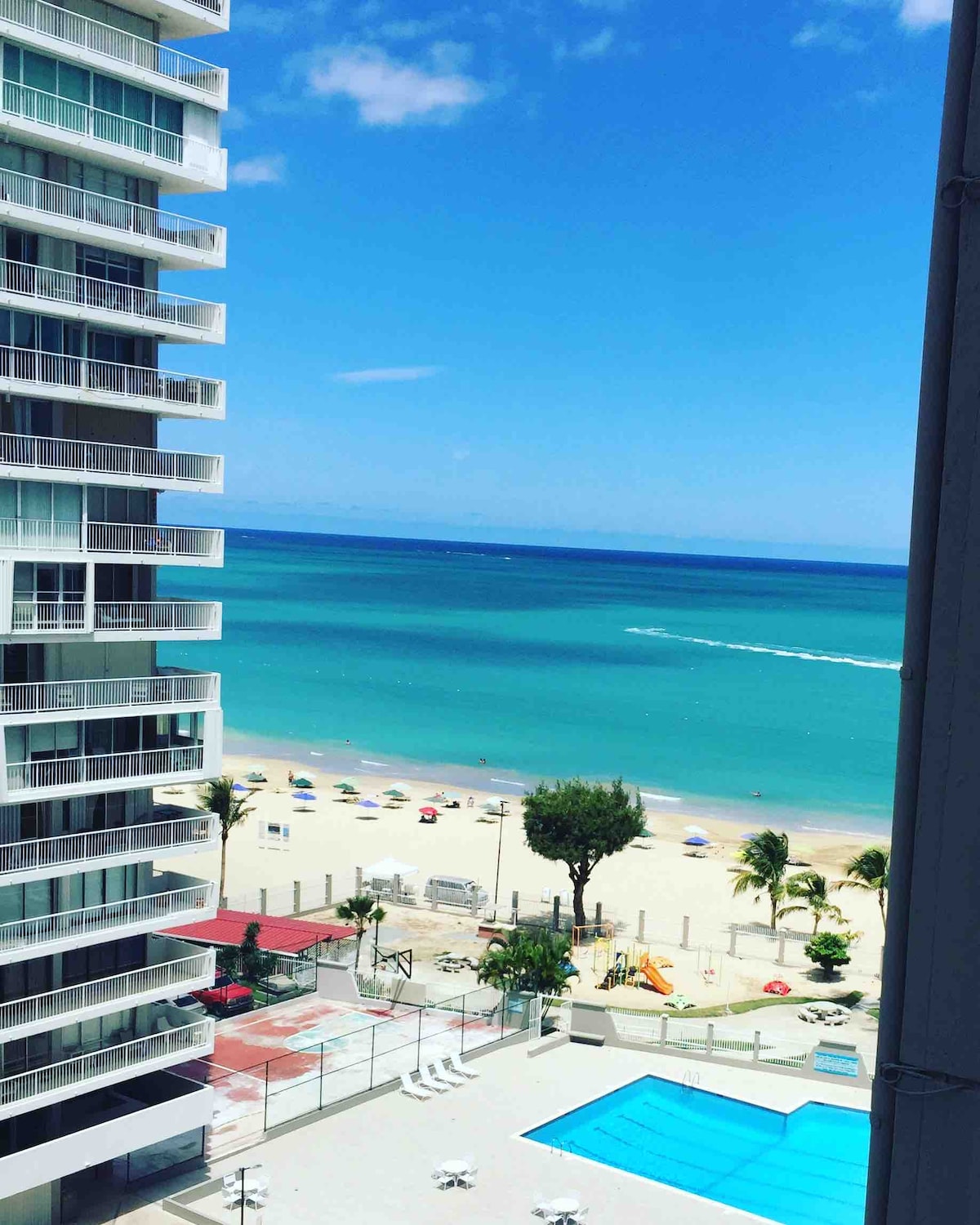 A panoramic view of a serene beach and turquoise waters is presented. The image shows a lively pool area with sun loungers, surrounded by trees and colorful umbrellas. Nearby, a sandy shore leads to the ocean, where gentle waves can be seen.