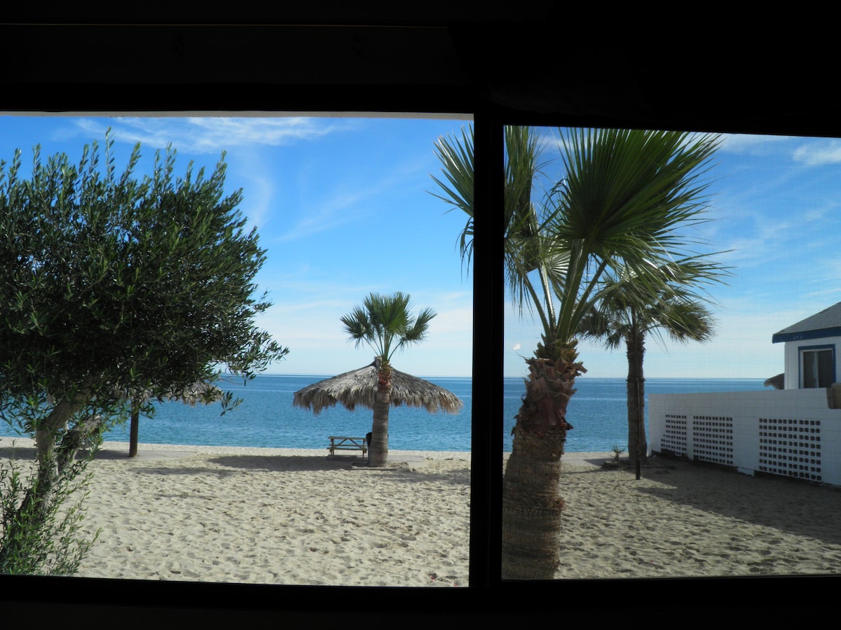 A view from the interior showcases a sandy beach framed by palm trees and a thatched umbrella. The tranquil sea extends to the horizon under a clear blue sky. A wooden picnic table is visible on the shore, offering a place for relaxation.