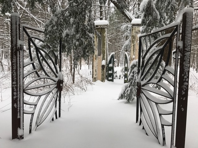 A whimsical gate, designed in the shape of butterfly wings, stands partially open, leading into a serene snow-covered garden. The path is blanketed in fresh snow, while trees and sculptures are softly draped in white, creating a tranquil winter scene.