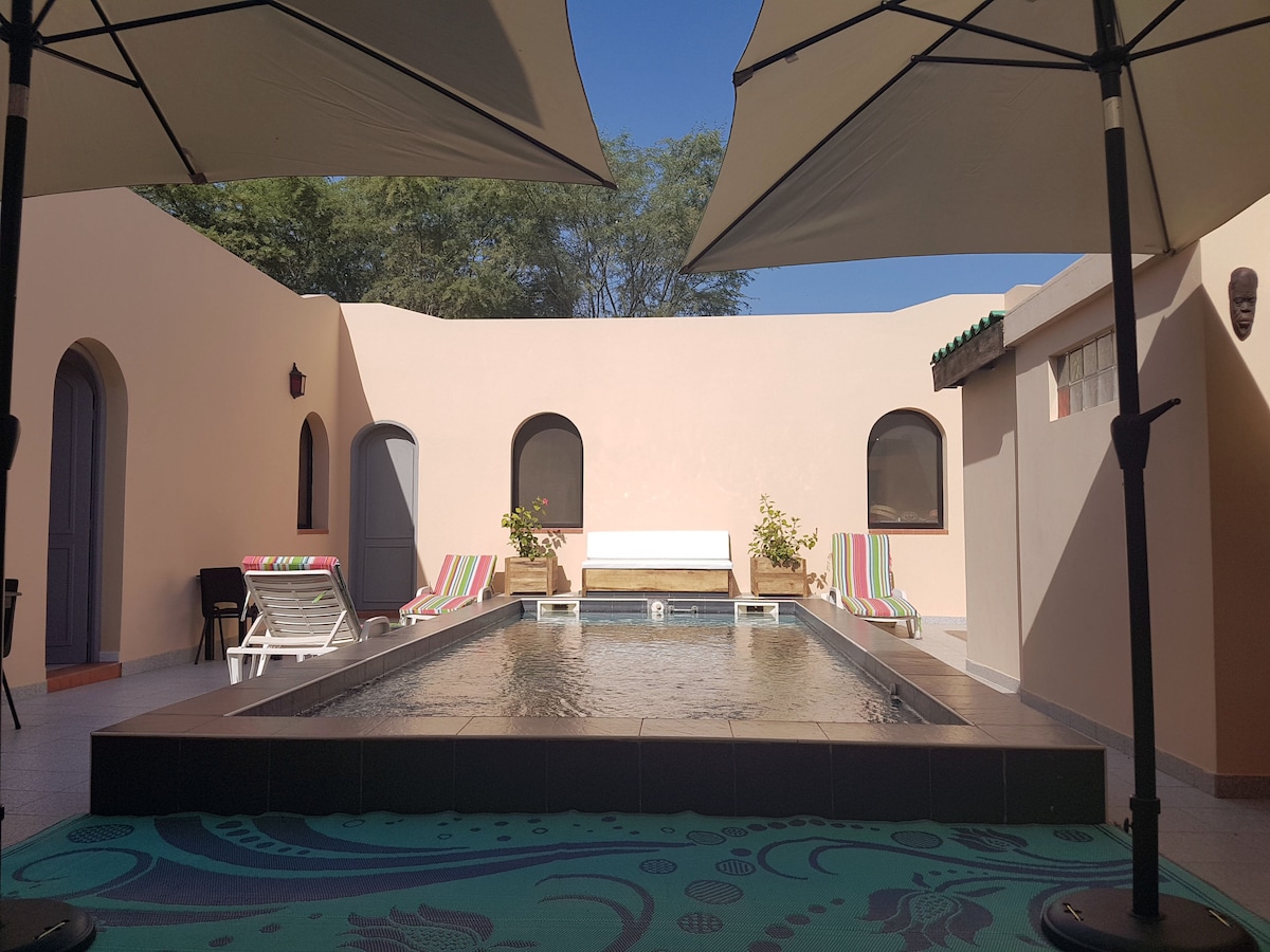 A private courtyard features a rectangular pool surrounded by sun loungers and colorful striped chairs. Umbrellas provide shade for relaxation, while plants add a touch of greenery. The walls are light-colored, enhancing the open atmosphere under clear blue skies.