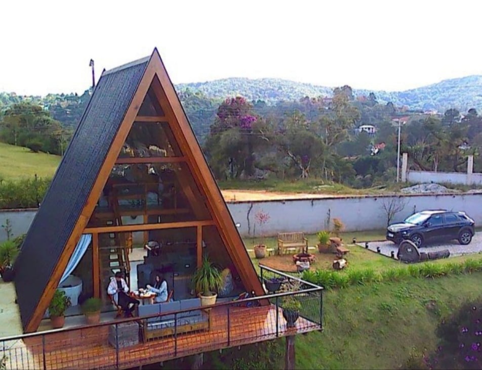 The modern A-frame structure features a spacious deck with outdoor seating and planters. A couple is seen enjoying a meal at a table, with expansive views of the surrounding nature and hills in the background. A vehicle is parked nearby.