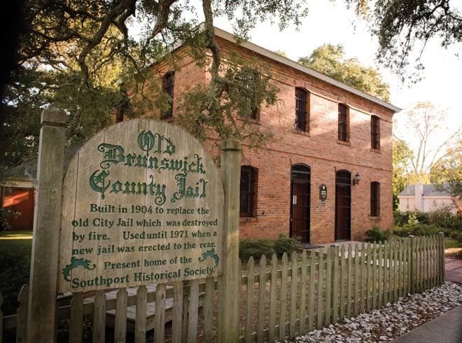 A historic brick building is framed by greenery and a wooden fence. A sign detailing the history of the Old Brunswick County Jail is prominently displayed in the foreground. The structure features arched windows and a central entry, surrounded by well-maintained landscaping.