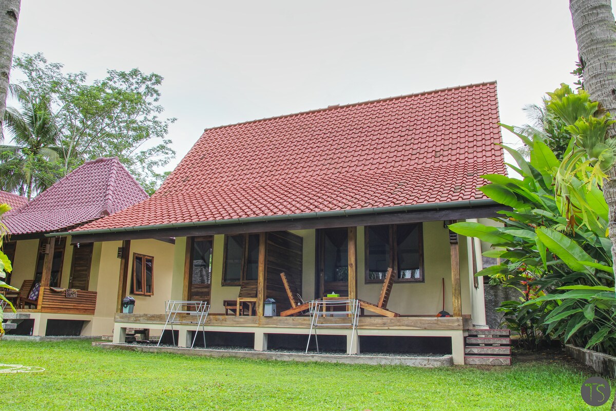 A charming house is surrounded by lush greenery and tall trees, featuring a sloped red tile roof. The porch presents several chairs, inviting a relaxed outdoor experience. Large windows allow natural light to fill the interior, providing a warm connection to the garden.