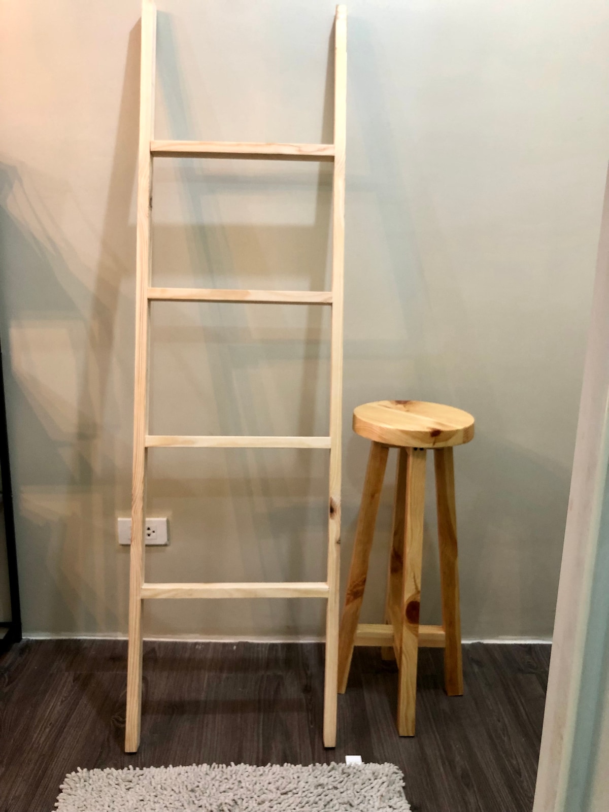 A wooden ladder with four rungs is positioned against a light-colored wall, creating a simple yet functional accent. Next to it, a round wooden stool with three legs offers additional seating. A small, soft rug sits at the base of the ladder, adding texture to the space.