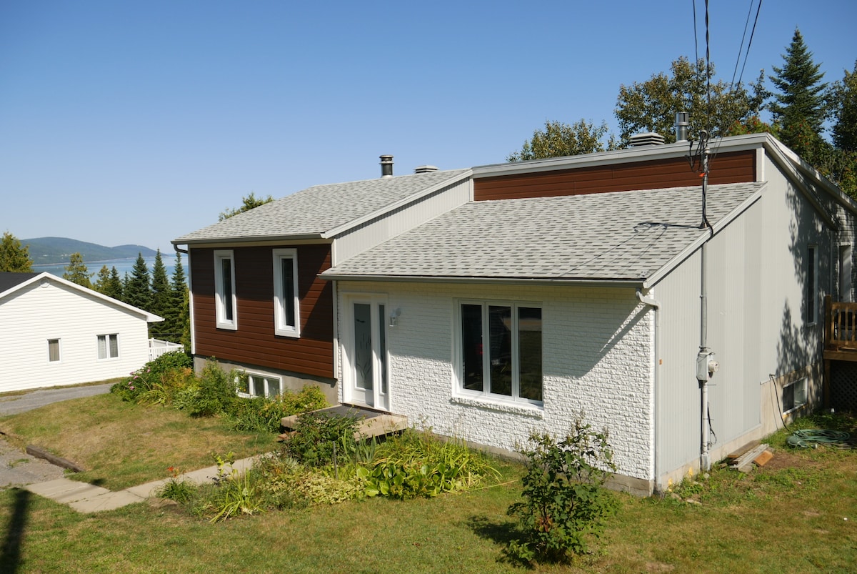 A spacious house is depicted with a combination of white and brown exterior walls. The entryway features a small porch with steps leading to the garden. Lush greenery surrounds the house, and the background showcases a hint of distant trees and a body of water.