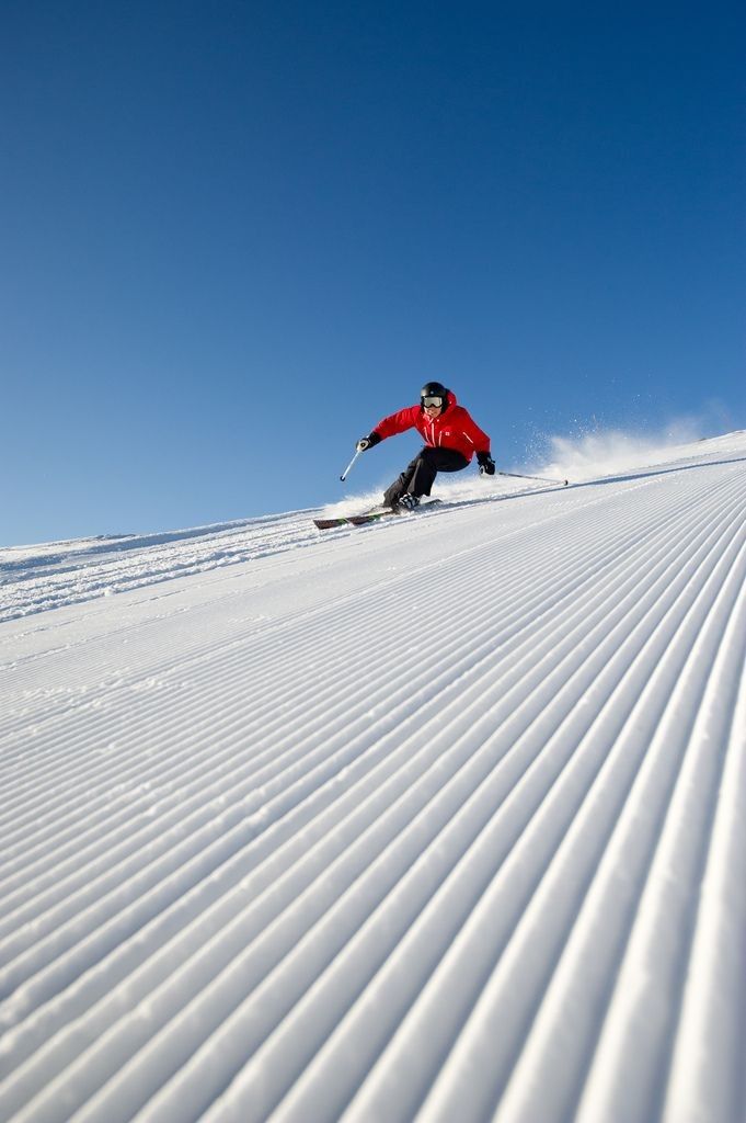 A skier is captured gliding down a freshly groomed slope, showcasing the smooth patterns of the snow beneath. The clear blue sky contrasts with the bright red attire of the skier, creating a dynamic scene representative of winter sports.