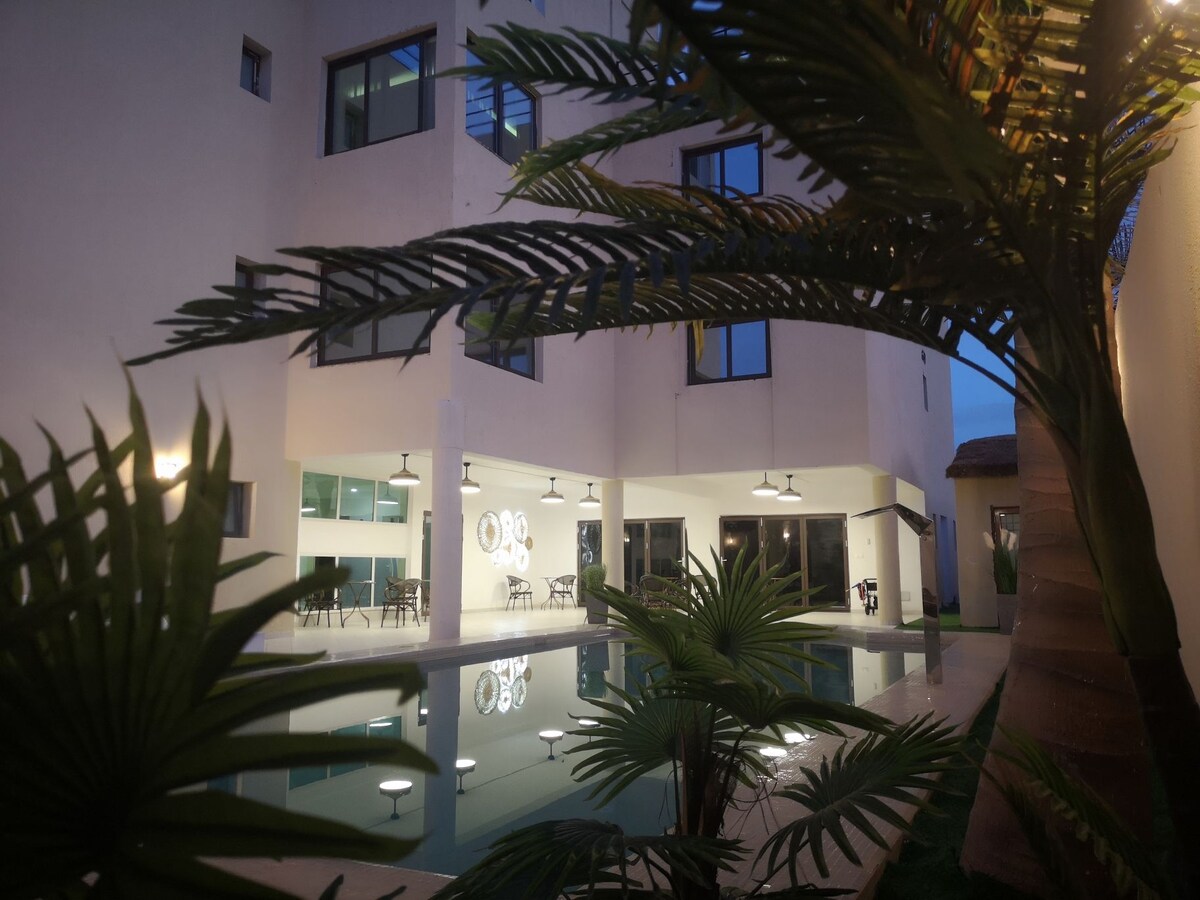 An inviting courtyard is framed by lush greenery, showcasing a serene pool surrounded by modern architecture. Soft lighting illuminates the area, creating a calm atmosphere, while large windows and a glimpse of outdoor seating can be seen in the background.