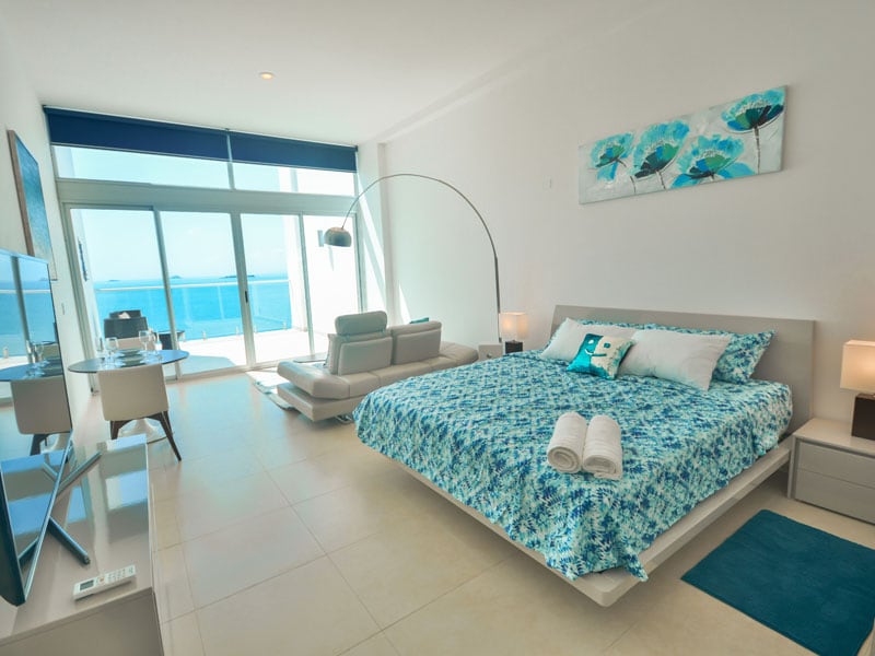 A spacious studio is equipped with a comfortable bed dressed in a vibrant blue pattern. A large window offers panoramic views of the ocean. Modern furniture includes a sleek sofa and a small dining area. Soft lighting emanates from bedside lamps, creating a serene ambiance.