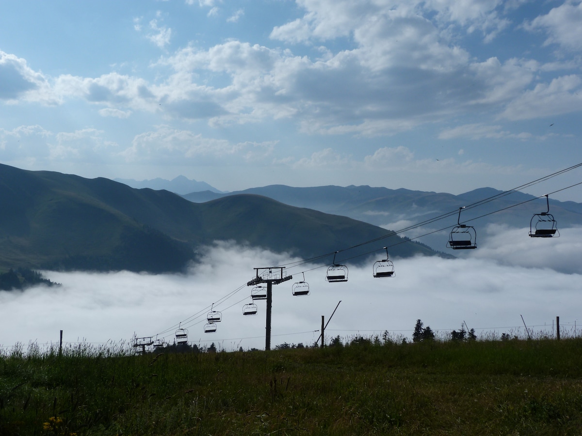 A ski lift is depicted ascending through a sea of clouds, surrounded by rolling green mountains. The horizon features misty peaks that blend into the sky. Grass and wildflowers are visible in the foreground, contributing to a serene landscape.