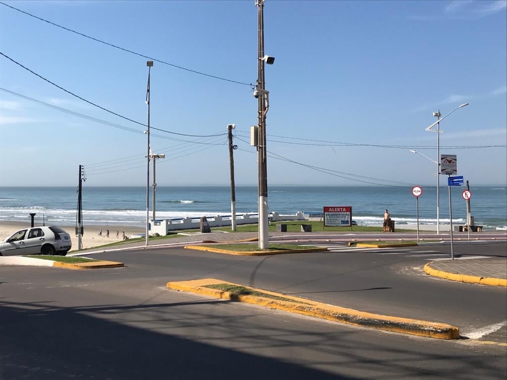 A view captures the intersection leading to the beach, with the ocean visible in the background. The scene includes a clear blue sky, a coastal road, and scattered palm trees, providing a sense of the location’s beachfront accessibility.