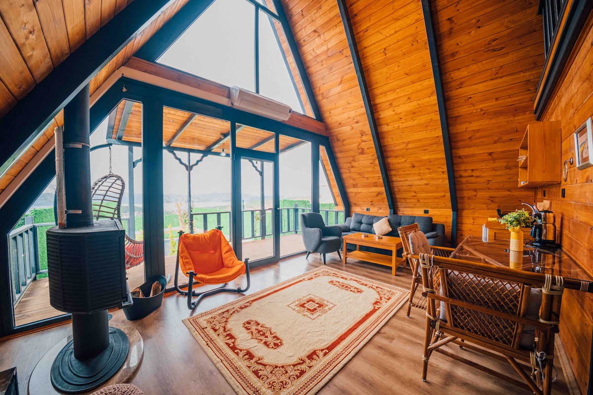 A bright loft area features high ceilings with wooden beams and large glass doors offering scenic views. Comfortable seating is arranged around a central rug, and a small dining area is visible near a kitchenette, enhancing the spacious and welcoming layout.