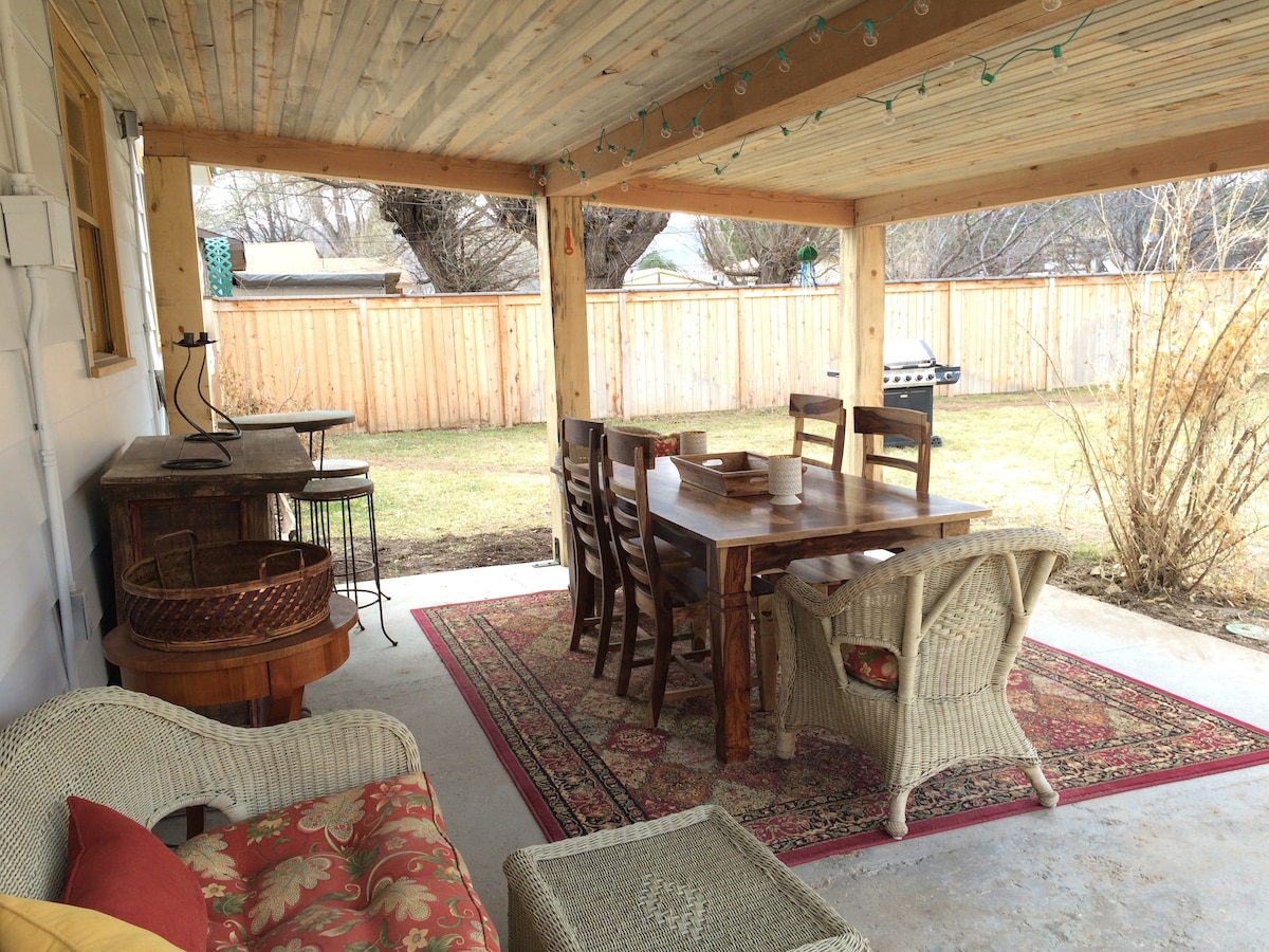 An outdoor patio area is covered and features a large dining table surrounded by wooden chairs. A cozy seating arrangement with wicker furniture and accents is positioned on a decorative rug. The fully enclosed yard provides privacy, with a grassy area visible beyond.