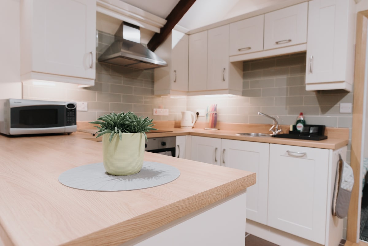 The kitchen area features ample countertop space with a small potted plant as a centerpiece. Modern cabinetry and appliances, including a microwave, are visible. Soft, ambient lighting enhances the overall functionality of the space, providing a warm and welcoming environment for cooking.