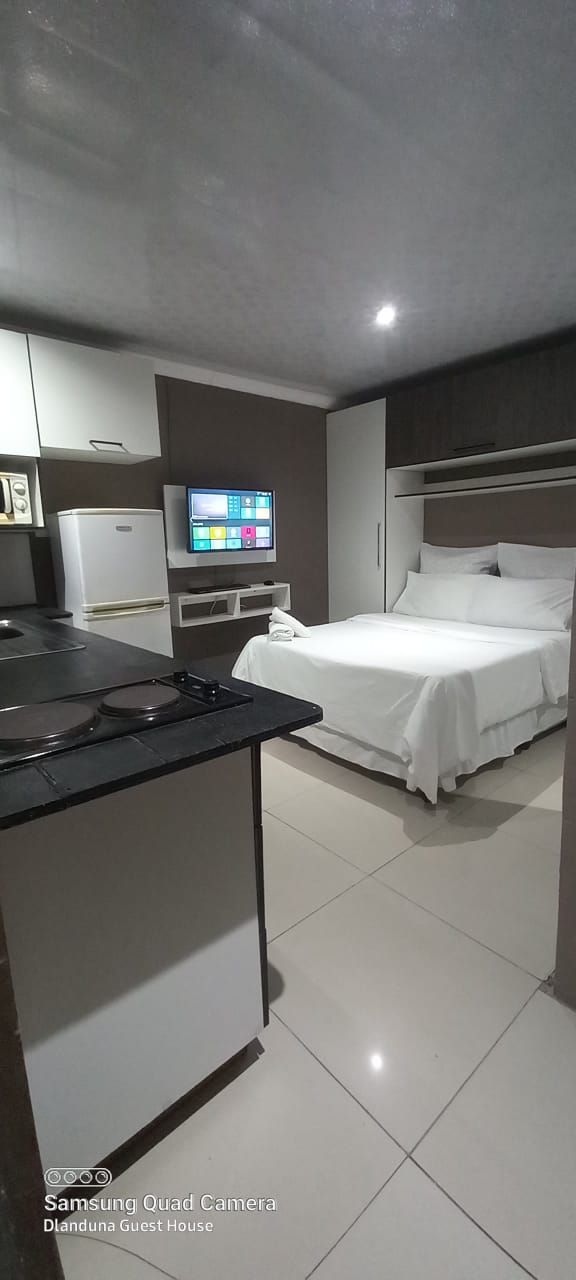 A modern studio apartment is featured with a compact kitchenette, equipped with essential appliances. A cozy bed with crisp white linens is positioned against the wall, and a wall-mounted flat-screen TV is visible. The space is bright, illuminated by soft ceiling lights, and tiled floors enhance the clean appearance.