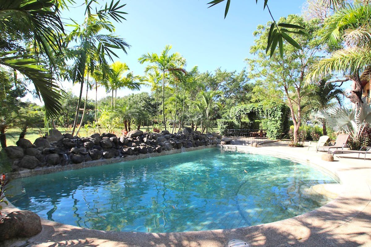 A tranquil pool area is surrounded by lush tropical landscaping, featuring palm trees and natural stone accents. The calm water reflects the blue sky, inviting relaxation. Comfortable lounge chairs are placed beside the pool, creating an ideal spot for sunbathing or unwinding.