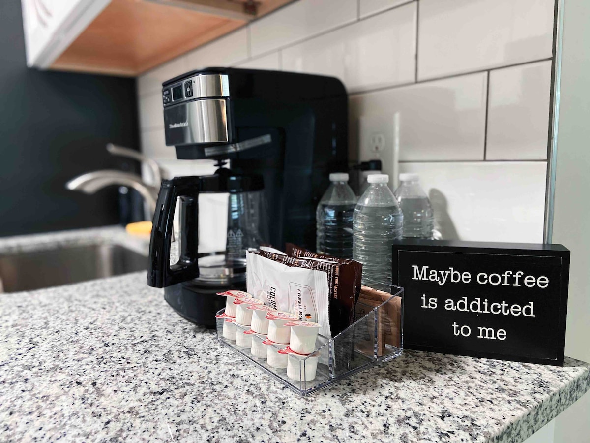 A coffee maker sits on a granite countertop beside a selection of coffee pods and water bottles. A sign with contrasting lettering states, 'Maybe coffee is addicted to me.' The modern kitchen backdrop features tiled walls and a stainless steel faucet.