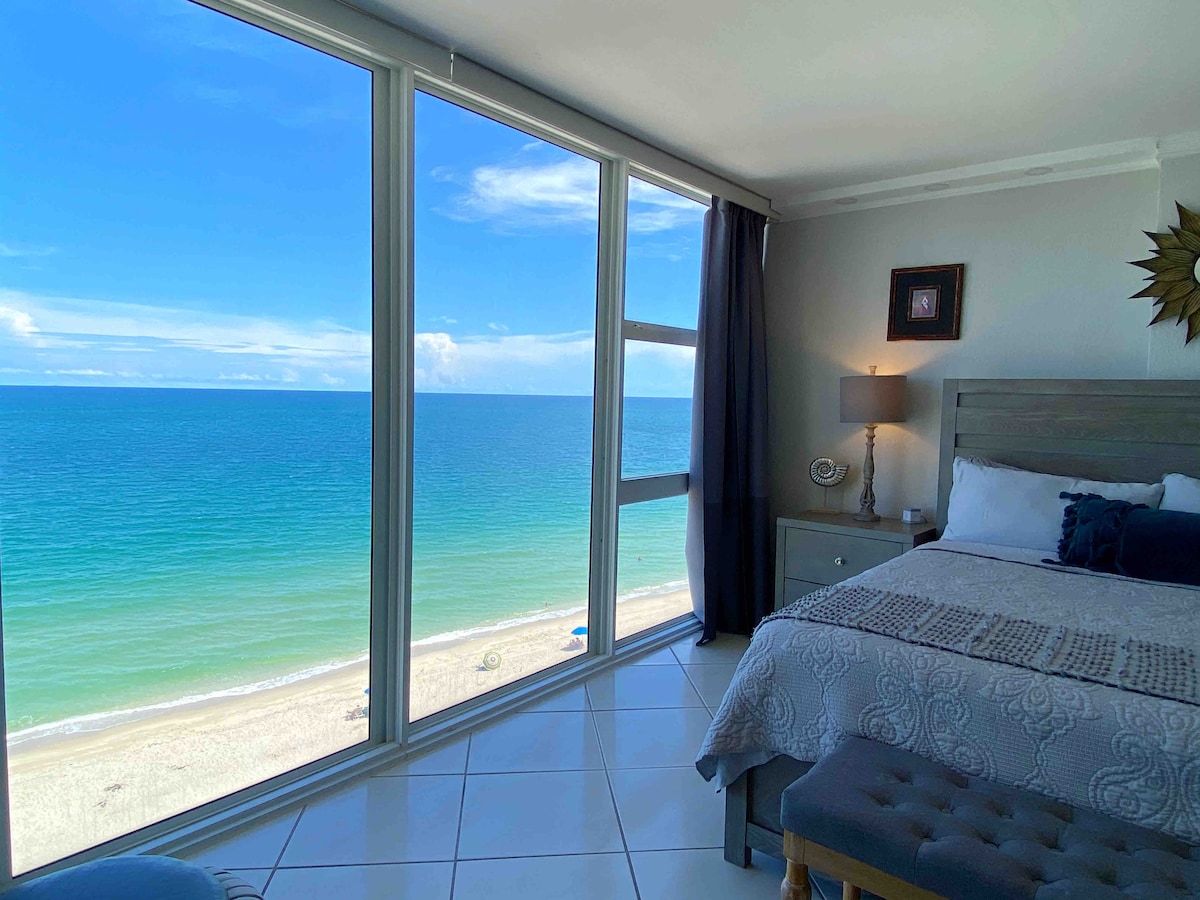 A serene bedroom features a spacious bed with neutral bedding. Large windows provide an unobstructed ocean view, showcasing the tranquil blue water and sandy beach below. Natural light fills the room, highlighting a bedside table and lamp, and a decorative wall piece adds a touch of elegance.