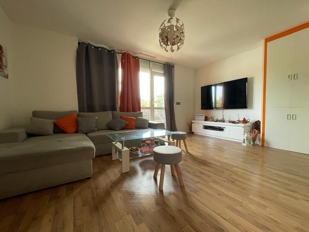 A living area features a comfortable gray sectional sofa with orange accent pillows. A glass coffee table sits on a warm wooden floor, surrounded by a small stool. Natural light enters through large windows adorned with sheer drapes, enhancing the spacious ambiance.