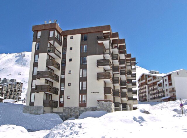 The exterior of the residence Le Prariond is presented, featuring a modern design with a combination of dark and light panels. Balconies are visible on the upper floors. Snow-covered ground surrounds the building, highlighting its location in a mountain setting.