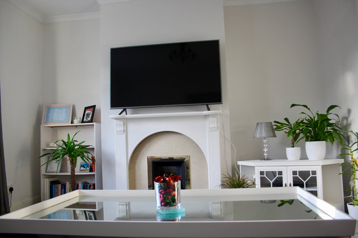A living area features a large wall-mounted TV above a decorative fireplace. A coffee table with a glass top sits in front, showcasing a small bowl of cherries. Plants are placed on both sides, adding greenery, while a white cabinet provides additional storage.