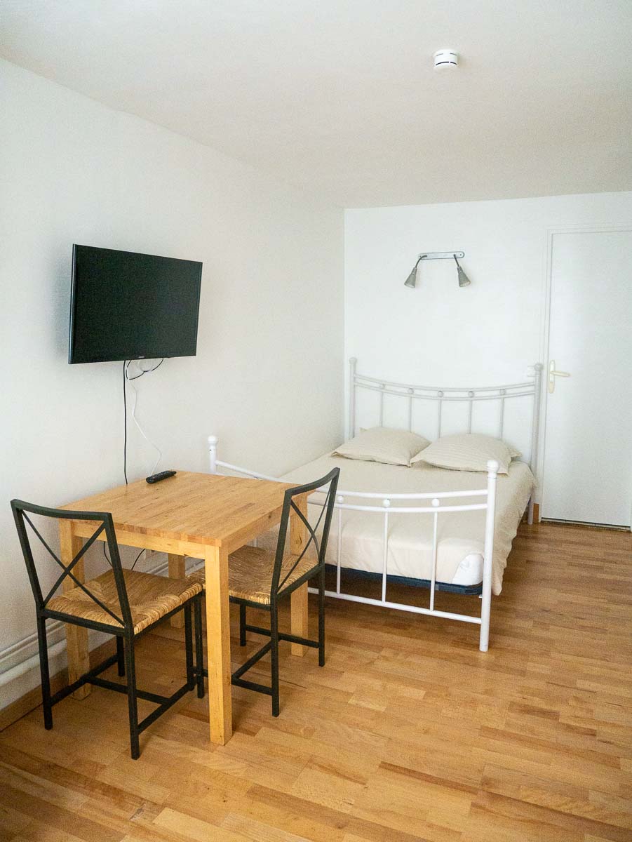 A compact studio features light wooden flooring and a minimalist design. A small wooden table paired with two chairs is visible beside a bed with a white frame. A wall-mounted TV is positioned across from the bed, creating a functional living space.