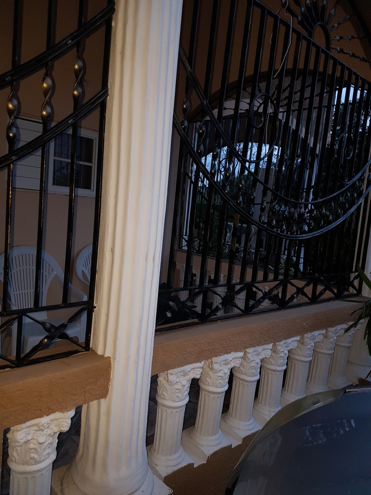 A front patio is visible, featuring decorated columns and a wrought iron railing. Two white chairs are positioned against the wall, providing a relaxing outdoor area. The surrounding greenery adds a touch of nature, enhancing the serene setting.