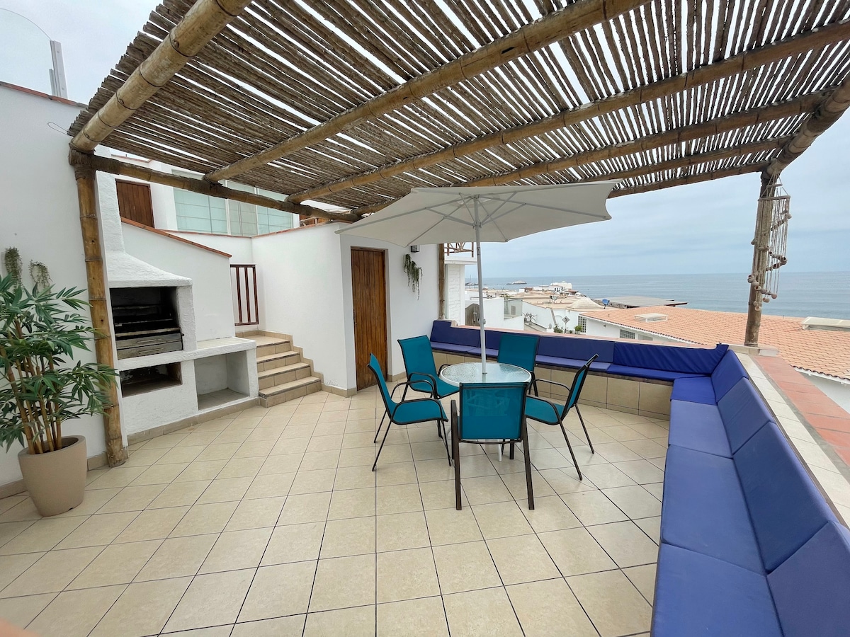 An expansive terrace is visible, featuring a round glass table surrounded by blue chairs. A covered area provides shade, and a built-in barbecue is situated against one wall, with an unobstructed view of the ocean in the background.