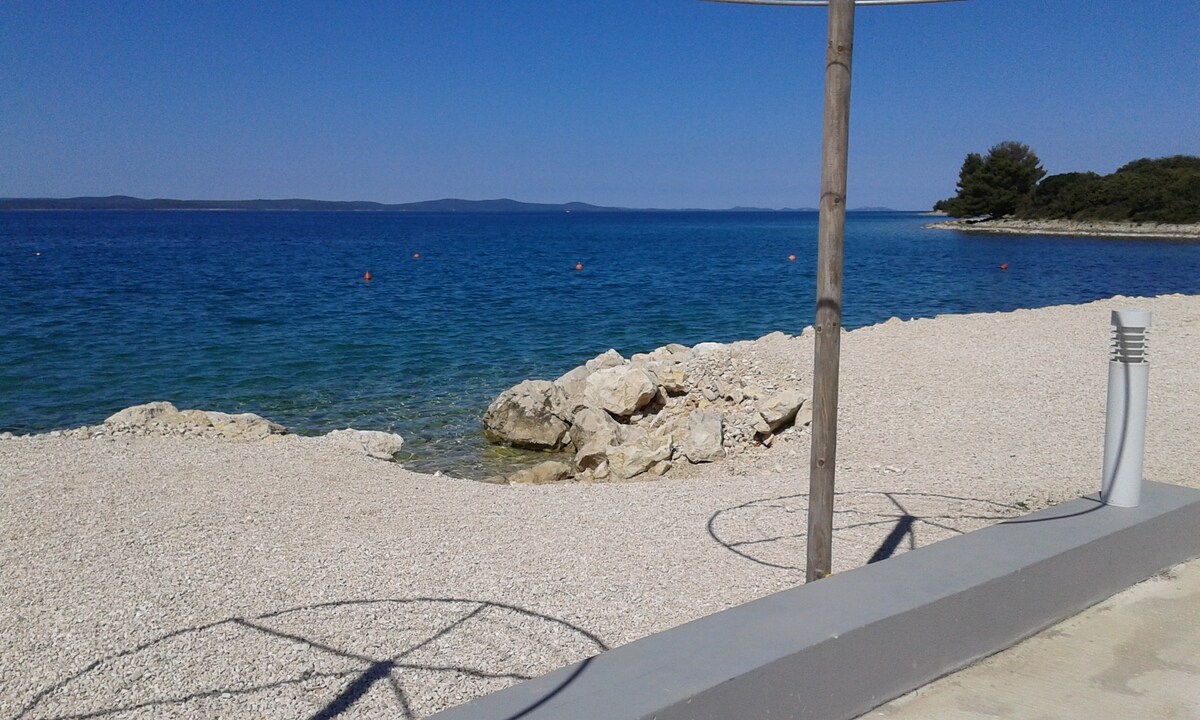 A clear view of the calm sea is presented, framed by a pebbled beach. Gentle waves lap against the shore, while distant rocky outcrops are visible in the water. A few trees line the coastline, and the bright blue sky enhances the serene atmosphere.