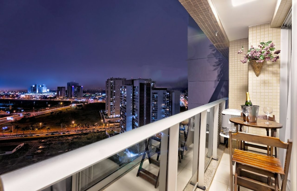 A small balcony is shown with elegant wooden furniture, including a dining table and two chairs. The city skyline is visible in the background, illuminated by nighttime lights. A bottle of champagne and two glasses are placed on the table, creating a refined outdoor setting.