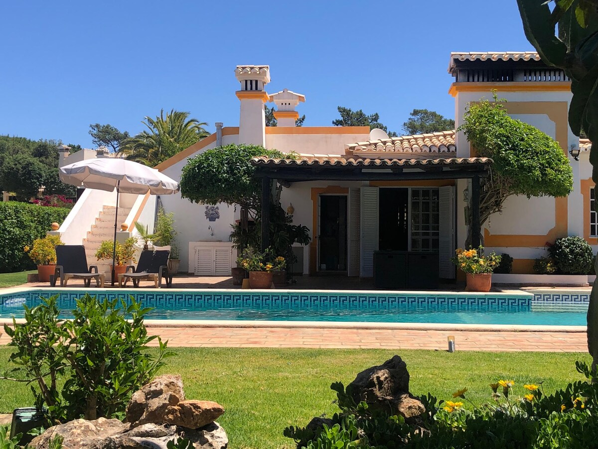 A spacious villa is featured, surrounded by lush greenery and a well-maintained garden. A private pool reflects the clear blue sky, while lounge chairs are positioned for relaxation. The exterior showcases traditional architecture, enhanced by vibrant potted plants and well-kept pathways.