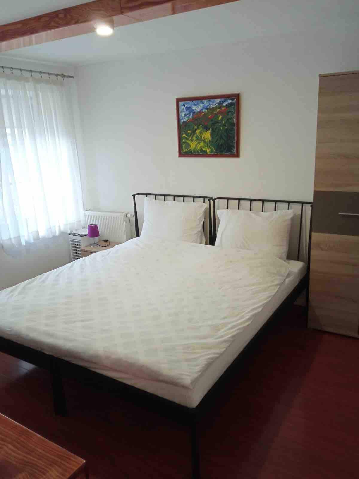 A cozy sleeping area features a spacious bed with white linens and two pillows. A colorful piece of wall art adds a touch of color. Natural light enters through a sheer curtain, highlighting the warm wooden floor and nearby wardrobe.