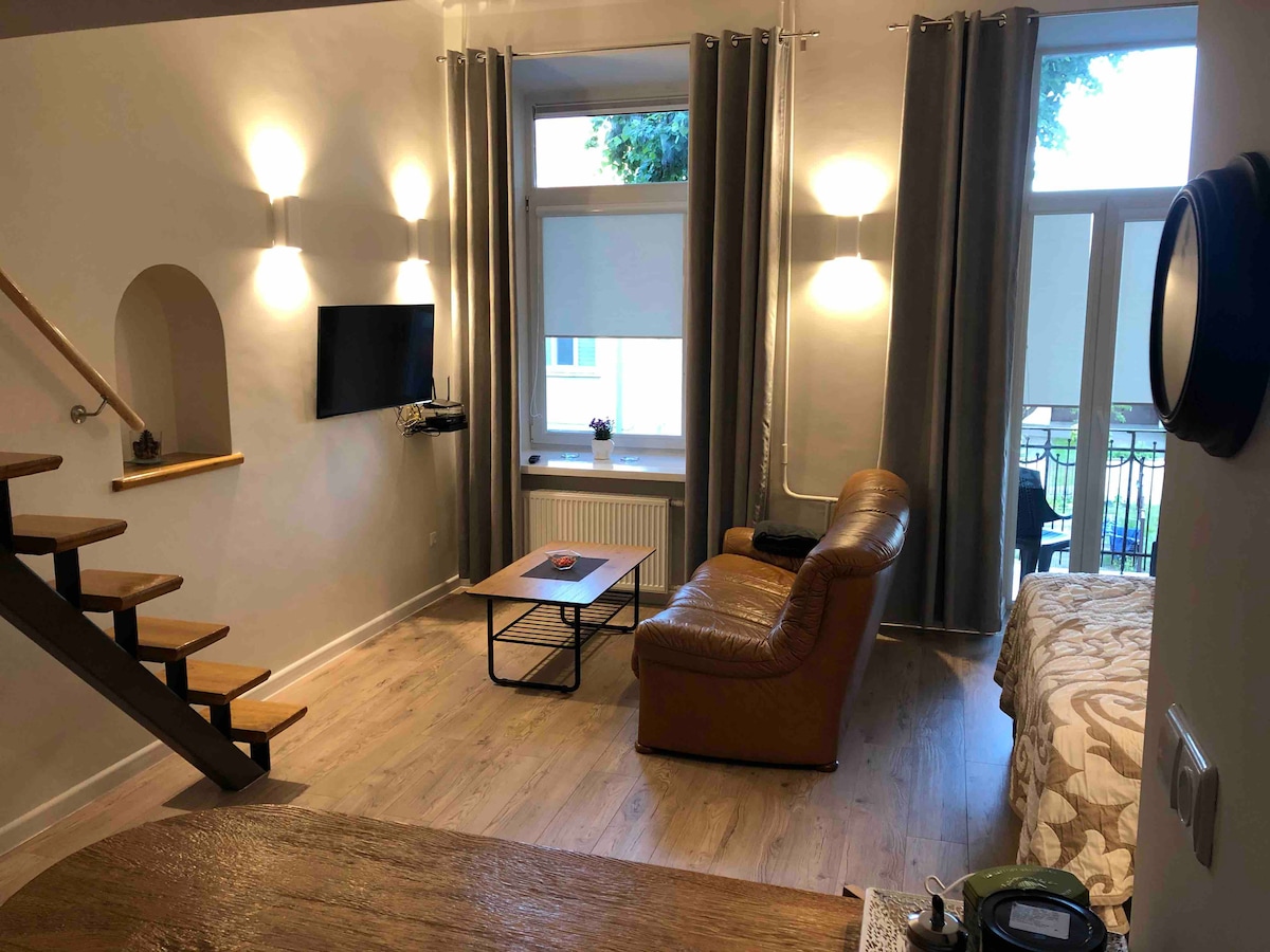 The living area features a comfortable leather sofa facing a wall-mounted TV, with soft lighting provided by adjacent wall sconces. A small coffee table is positioned in front of the sofa. A staircase leads to the upper level. Large windows allow natural light and provide access to a balcony.