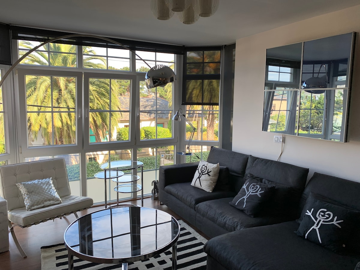 The living room features a spacious black sofa adorned with decorative cushions. A round glass coffee table is centered on a striped rug. Large windows allow natural light to fill the space, with views of palm trees visible outside.