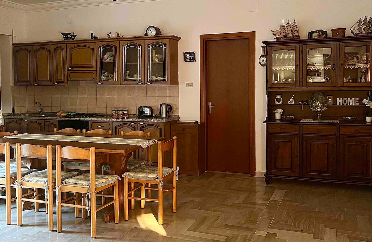 An open plan kitchen and dining area features wooden cabinetry with glass doors, a spacious dining table surrounded by six chairs, and various kitchen appliances. A dark wood sideboard is present, displaying decorative items, including nautical motifs, creating a warm and inviting space.