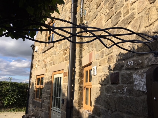 The image captures the exterior of a stone cottage, highlighting the textured stone walls and wooden window frames. The back door is visible, accompanied by utility connections. A lush green hedge and a cloudy sky fill the backdrop, adding to the rural charm of the setting.