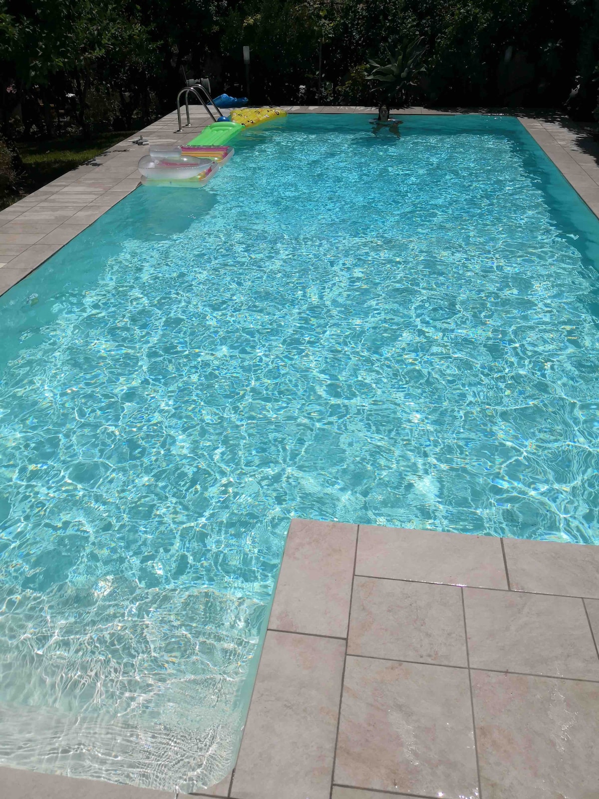 A private swimming pool is featured, with clear turquoise water reflecting sunlight. The surrounding area is paved with large tiles, and colorful inflatable floats are visible at the pool's edge. Lush greenery is present in the background, offering a sense of tranquility.