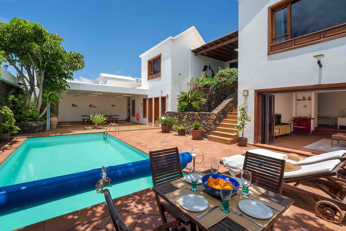 A well-maintained outdoor area features a private swimming pool surrounded by tiled flooring and potted plants. A dining table for four is set with glasses and a fruit bowl, with sun loungers positioned nearby. The villa's walls are bright, and a staircase leads to an upper level.