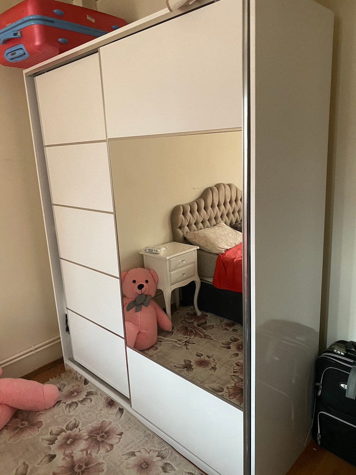 A white wardrobe with mirrored sliding doors is positioned against the wall. A plush pink teddy bear is placed on the carpet nearby. A cozy bed with a tufted headboard is visible in the background, along with a small bedside table.