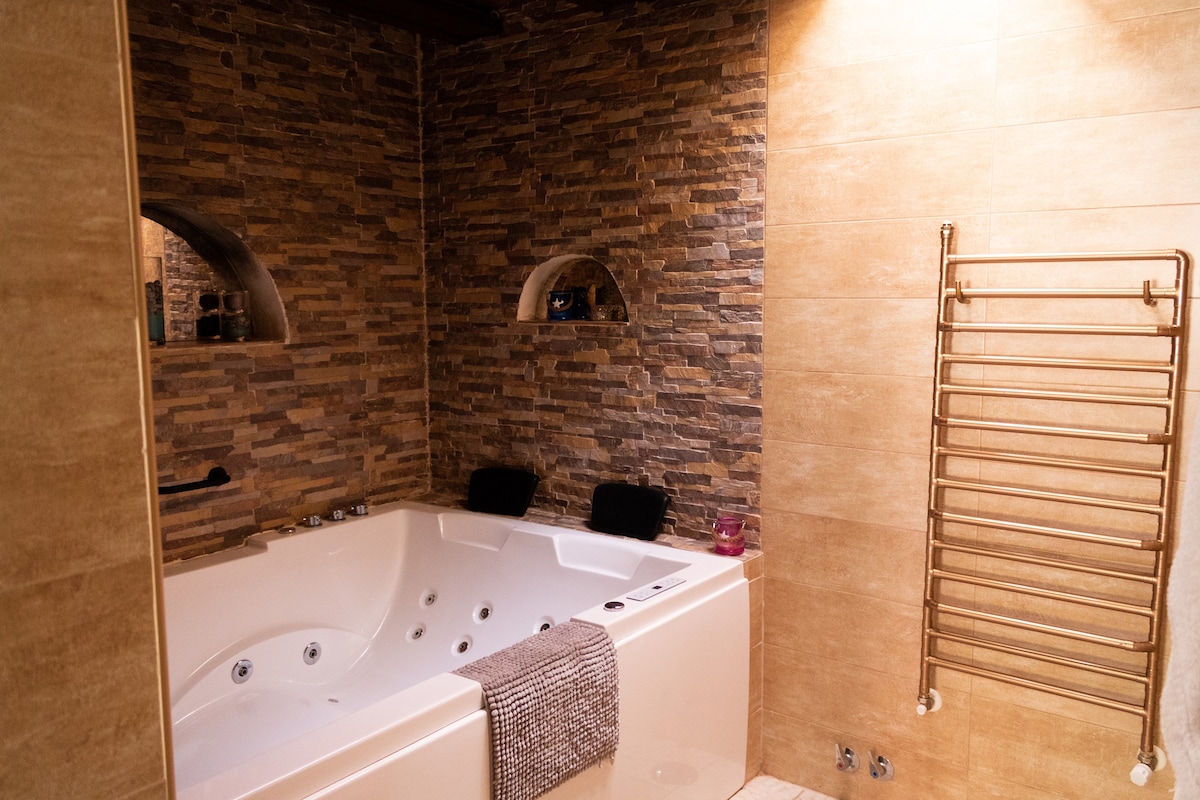 A spacious bathroom features a rectangular jacuzzi surrounded by textured stone walls. Soft towels are neatly placed on the edge of the tub, and a towel warmer is mounted on the wall, providing a blend of luxury and comfort.