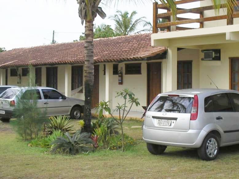 A serene outdoor view of a property featuring a row of suites with terracotta roofs. Vehicles are parked nearby on a grassy area, surrounded by tropical plants and palm trees, contributing to a relaxed atmosphere.