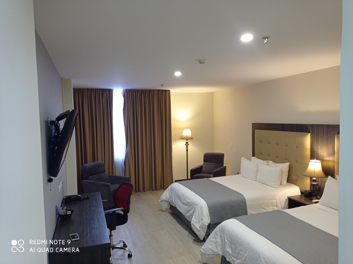 A well-appointed guest room features two spacious beds with crisp white linens. A desk with a chair is positioned next to a large television mounted on the wall. Soft lighting is provided by a lamp on the side table, and curtains frame the window, allowing natural light to enter.