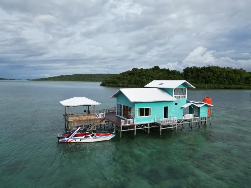 A striking stilted house with a vibrant turquoise exterior is situated over clear water. Nearby, a small boat is tethered at the dock. Lush greenery surrounds the property, showcasing a serene coastal environment under a cloudy sky.