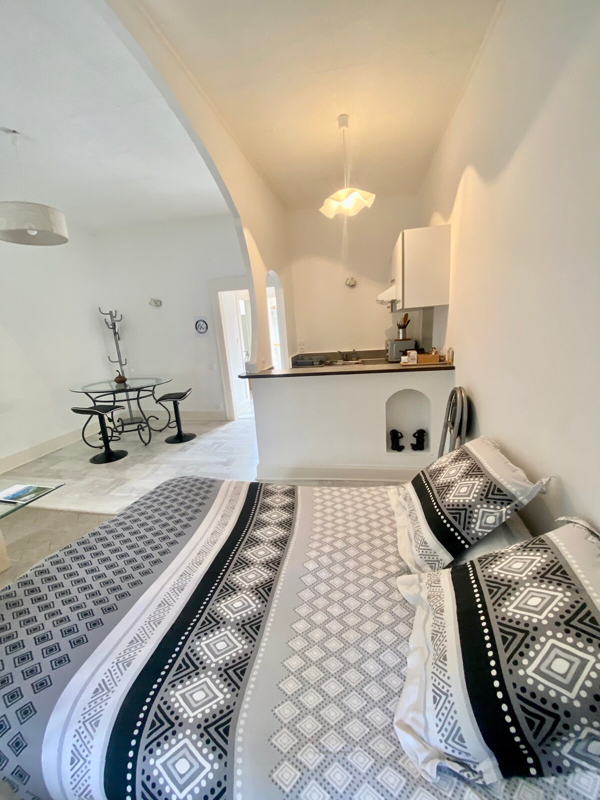 The studio features a cozy sleeping area with a patterned bedspread, complemented by neutral walls. A small dining table with chairs is visible beside the kitchenette, which includes essential appliances. Natural light is invited through the nearby doorway, enhancing the bright overall atmosphere.