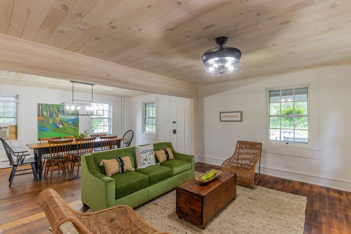 A spacious living area is highlighted by a green sofa and a wooden coffee table, complemented by rattan chairs. Natural light fills the room through multiple windows, showcasing a stylish dining space in the background with a wood table and black chairs.