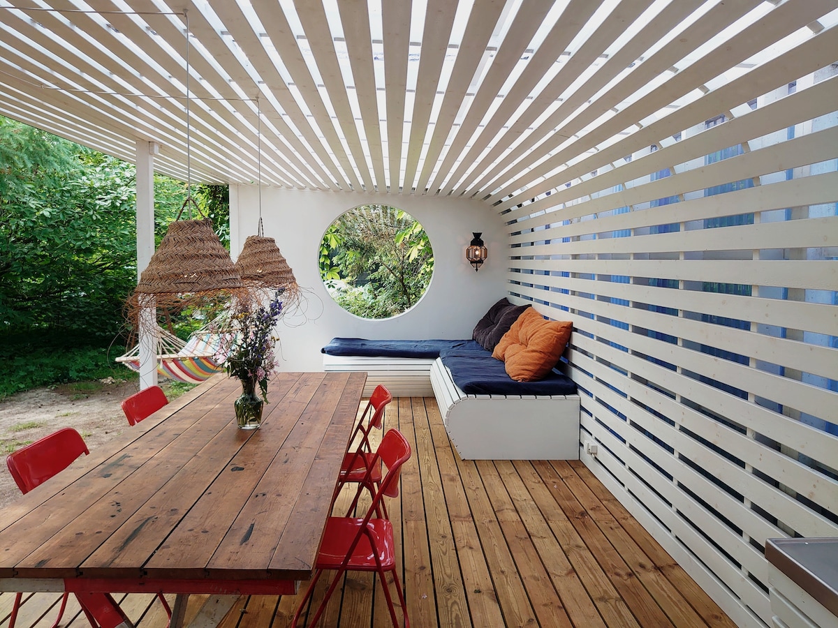 A bright seating area is defined by a striking circular window and a long wooden table surrounded by red chairs. A cozy bench with cushions offers additional seating. The space is framed by horizontal slats, allowing glimpses of the lush greenery outside.