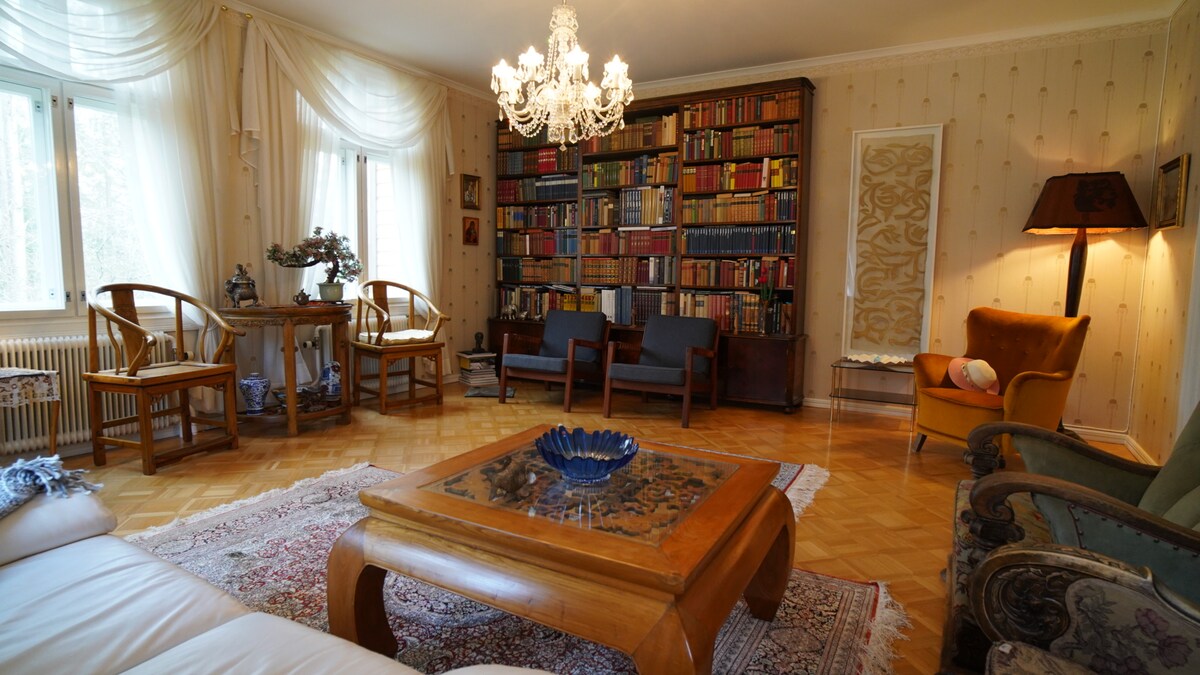 A spacious living room features a decorative chandelier above a wooden coffee table. Surrounding furniture includes a mix of armchairs and sofas, positioned near a large bookshelf filled with books. Natural light enters through the windows, enhancing the room's welcoming ambiance.