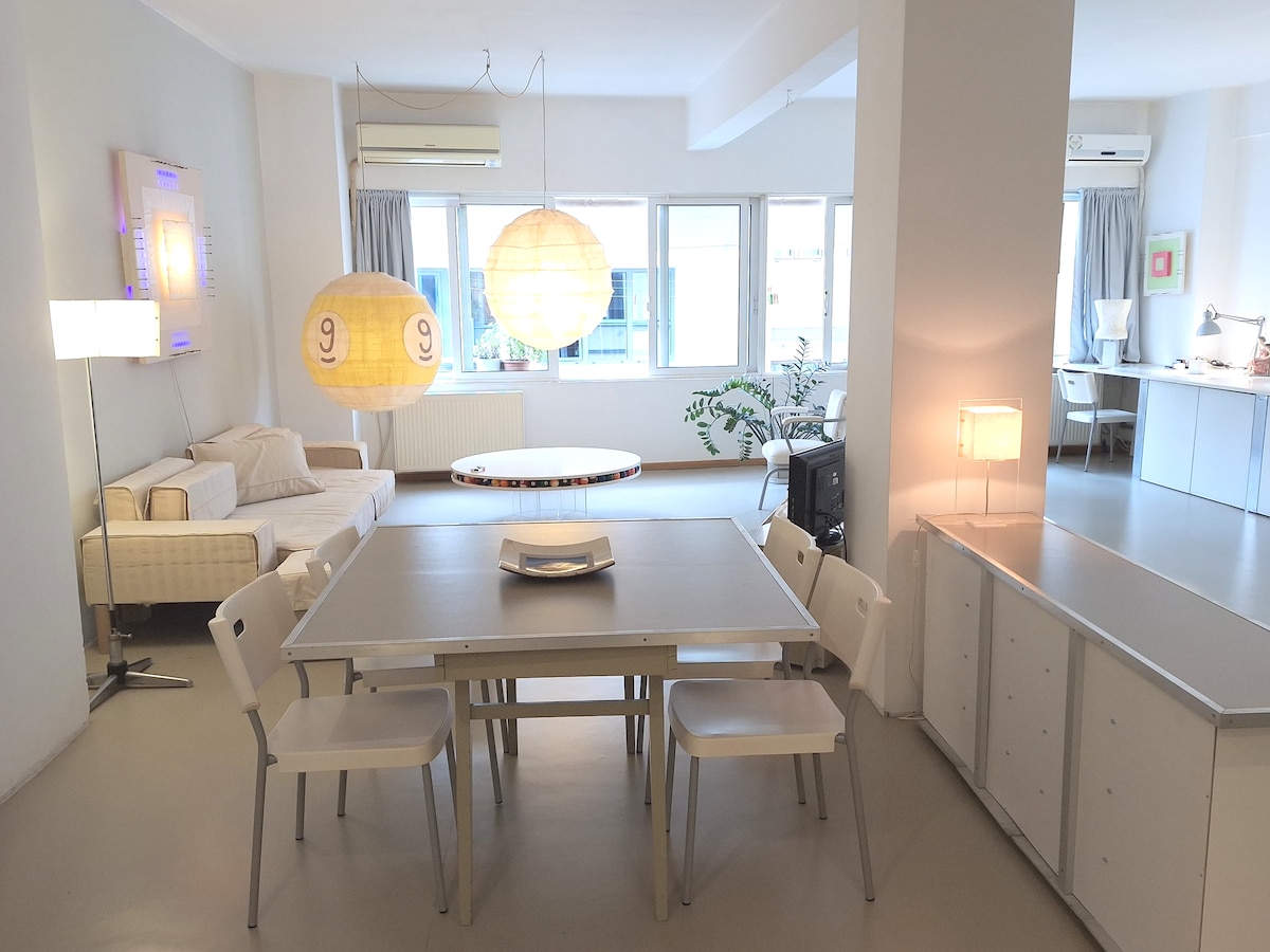 An open-plan living area features a dining table surrounded by white chairs, with a large circular table in the center. Soft seating is located to the left, illuminated by large pendant lights. Windows provide natural light, enhancing the spacious layout.