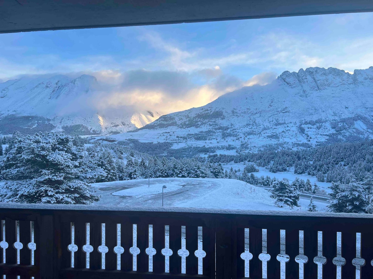 A snow-covered landscape is visible from the balcony, with majestic mountains in the distance partially shrouded in clouds. The ground is blanketed in fresh snow, and a serene valley can be seen, creating a peaceful winter scene.